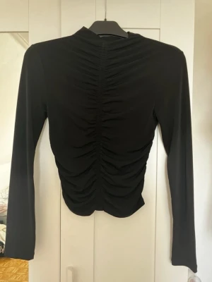 Svart långärmad topp med rynk - Trendig svart långärmad topp med rynkdetaljer längs hela framsidan och baksidan. Hög hals och figurnära passform ger en snygg siluett. Perfekt att styla med jeans eller kjol för en cool look.