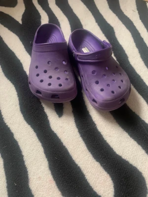 Lila tofflor i crocs-stil - Säljer ett par lila tofflor i klassisk crocs-stil med ventilerande hål och justerbar hälrem. Tillverkade i lätt och bekvämt plastmaterial, perfekta för att glida i hemma eller ute. Färgen är en snygg lila nyans och designen är enkel men trendig.