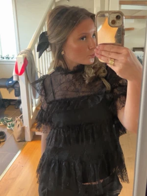 Svart spetsblus med volanger - Säljer en svart blus i genombruten spets med flera lager volanger och låg hals. Blusen har korta ärmar med volangdetaljer och är transparent, perfekt till fest eller vanlig outfit. Supersnygg och trendig topp från Malina, originalpris 1800kr.