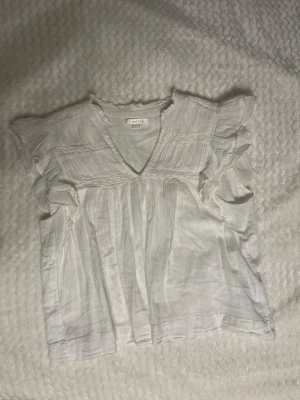 Vit blus - Söt vit blus från Gina tricot med v-ringning och volangdetaljer vid axlarna. Blusen har korta ärmar och är tillverkad i ett lätt, luftigt tyg som känns perfekt för sommaren. Smockad upptill och lös passform för en avslappnad look. 