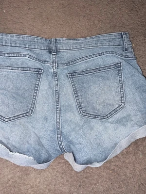 Blå jeansshorts med rå kant - Säljer ett par klassiska blå jeansshorts med rå kant nertill och två bakfickor. Shortsen har en avslappnad passform och är perfekta för varma dagar. Tillverkade i bomull med lite polyester och elastan för extra komfort.