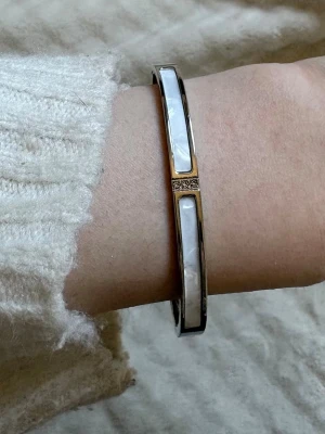 Silver armband med pärlemordetaljer - Snyggt armband i silverton med vita pärlemordetaljer och små glittrande stenar i mitten. Armbandet har en stilren och modern design med raka linjer och passar perfekt för dig som gillar minimalistiska accessoarer.