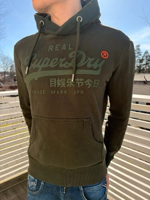 Mörkgrön Superdry hoodie med tryck - Snygg mörkgrön hoodie från Superdry med stort logotryck och japanska tecken på bröstet. Hoodien har snörning i huvan, känguruficka och ribbade muddar. Perfekt för en avslappnad och cool stil. Det går inte riktigt att se den riktiga färgen på bild men i verkligheten är den mer grön. Hör gärna av dig vid minsta fundering!