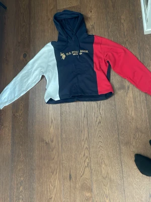 Flerfärgad hoodie från U.S. Polo Assn. - Snygg hoodie från U.S. Polo Assn. i färgblock med marinblå, vit och röd. Guldigt tryck och logga på bröstet. Klassisk huva och långärmad modell, perfekt för en avslappnad streetwear-look. Mjuk och bekväm bomullsblandning.