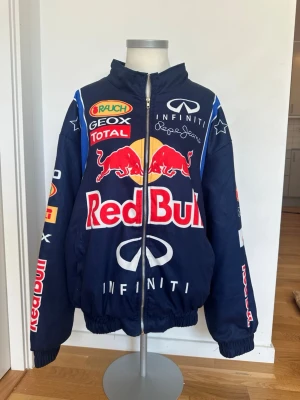 Red Bull blå bomberjacka - Blå bomberjacka från Red Bull med stora tryckta loggor och patchar på fram- och baksida samt ärmar. Jackan har dragkedja, hög krage och elastiska muddar. Perfekt för dig som gillar motorsport och streetwear.