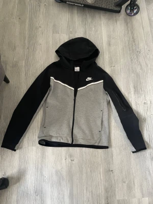 Nike tech hoodie i svart och grå - Nike hoodie med dragkedja i svart och grått. Snygg vit detalj över bröstet och klassisk Nike-logga på vänster sida. Huva och långa ärmar, samt en praktisk ficka med dragkedja på ena ärmen. Tillverkad i mjukt sweatshirtmaterial, perfekt för en sportig look.