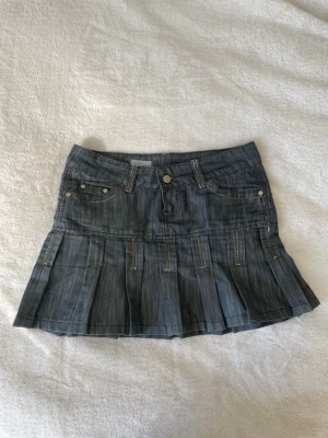 Kort jeanskjol med veckad nederdel - Säljer en snygg kort grå jeanskjol med veckad nederdel och klassiska fickor fram och bak. Kjolen har bälteshällor, knapp och dragkedja framtill samt dekorativa sömmar. Perfekt för en trendig och avslappnad look.