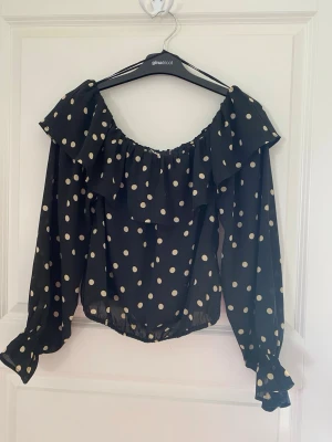 Prickig offshoulder blus från BikBok - Snygg svart offshoulder blus från BikBok med beige prickar. Blusen har volang vid axlarna, långa ärmar med resår vid handlederna och en luftig passform. Tillverkad i ett lätt och skönt material som ger en trendig look. Kolla gärna mina andra annonser❗️