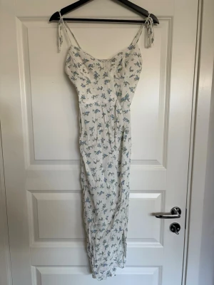 Vit långklänning med blå blommor - Söt långklänning med smala axelband som knyts på axlarna och en slits. Klänningen är vit med blått blommönster och har en figurnära passform. Köpt på Bershka i storlek M och endast använd en gång på midsommar därav mycket bra skick. Perfekt för somriga tillfällen🤗