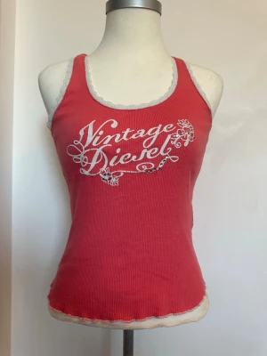 Rosa ribbad linnetopp från Diesel - Säljer en rosa ribbad linnetopp från Diesel med vit spetskant och trycket 'Vintage Diesel' i vitt framtill. Toppen är ärmlös och har en figurnära passform med snygga detaljer som ger en retrovibe.