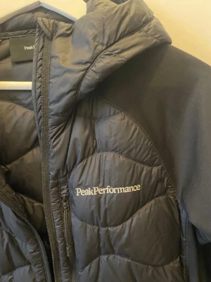 Svart pufferjacka Peak Performance - Snygg svart pufferjacka från Peak Performance med huva och dragkedja. Jackan har vadderade partier och släta ärmar med Peak Performance-logga på bröstet och ärmen. Perfekt för kalla vinterdagar och riktigt skön att bära.