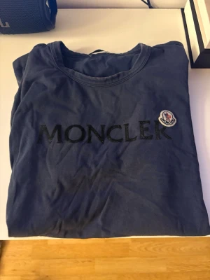 Blå Moncler t-shirt med logga - Snygg mörkblå t-shirt från Moncler med stor svart logga på bröstet och det klassiska Moncler-märket på vänster sida. T-shirten har rund hals och är tillverkad i mjuk bomull för en skön känsla. NFC Scan funkar!