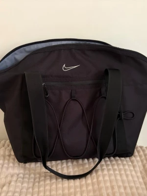 Svart axelbandväska från Nike - Snygg svart axelbandväska från Nike med klassisk vit logga framtill. Väskan har breda axelband, dragkedja upptill och en praktisk ytterficka med elastiska detaljer. Tillverkad i slitstarkt tyg, perfekt för skolan eller gymmet.