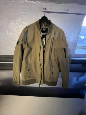 Beige bomberjacka från Superdry - Snygg beige bomberjacka från Superdry med camofodrad insida och ribbade muddar. Jackan har dragkedja, fickor med knappstängning och en ärmficka med dragkedja. Märkespatch på ärmen och coola detaljer på insidan.