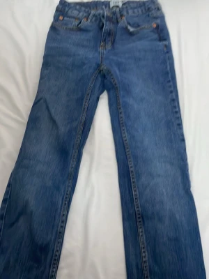 Blå raka jeans med hög midja - Klassiska blå jeans med rak passform och hög midja. Jeansen har fem fickor, bälteshällor och knappgylf. Materialet är kraftig denim i bomull. Perfekta för en avslappnad och tidlös stil.