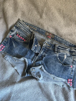 Blå jeansshorts med rosa nitar från only  - Säljer ett par blå jeansshorts från Only med råa kanter och slitningar. Shortsen har coola rosa nitar vid fickorna och en klassisk femficksmodell. Perfekta för sommaren och ger en edgy vibe till din outfit.