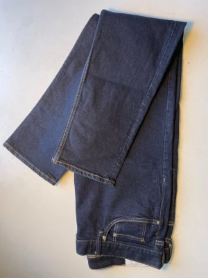 Mörkblå raka jeans med kontrastsömmar - Säljer ett par mörkblå jeans med raka ben och klassiska kontrastsömmar. Jeansen har fem fickor, bälteshällor och stängs med knapp och dragkedja. Materialet är kraftig denim som ger en tidlös look.