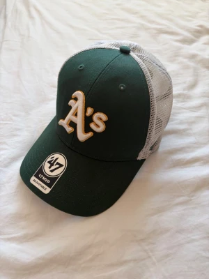 Grön Oakland A's keps från 47 Brand - Snygg grön keps med vit mesh baktill och broderad Oakland A's-logga i gult och vitt framtill. Justerbar passform med klassisk böjd skärm. Perfekt för dig som gillar sportig stil och vill ha en luftig keps till sommaren.