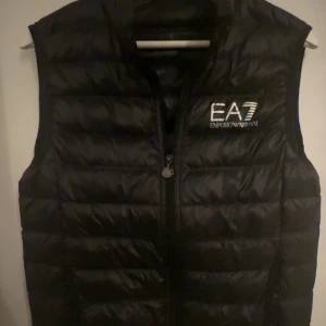Svart dunväst från Emporio Armani - Snygg svart dunväst från Emporio Armani med EA7-logga på bröstet.         STORLEK S                 Västen har en dragkedja framtill och är i perfekt skick inte trasig någonstans. Original pris (1999kr ) mitt pris (400). Men pris kan diskuteras. Och byten kan jag ta också.💸🍾🥂