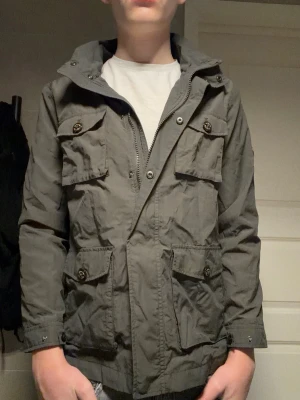 Mörkgrå vindjacka med fickor - Stilren mörkgrå vindjacka från Massimo Dutti med fyra stora fickor framtill och dragkedja. Jag är ca 175 cm så jag skulle nog säga att den passar bra för dem som är runt 170 cm. Tillverkad i ett lätt, vindtätt material som passar perfekt för blåsiga dagar.