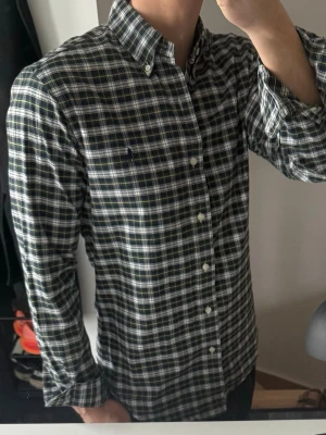 Rutig skjorta från Polo Ralph Lauren - Klassisk rutig skjorta från Polo Ralph Lauren i mörkblått, vitt och grönt. Skjortan har lång ärm, button-down krage och broderad logga på bröstet. Tillverkad i mjuk bomull och har normal passform. Perfekt för en avslappnad men ändå stilren look.