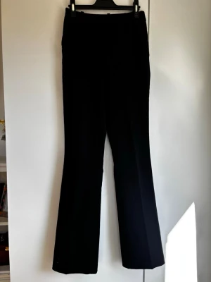 Svarta utsvängda kostymbyxor - Säljer ett par svarta kostymbyxor med utsvängda ben och hög midja. Byxorna har en klassisk, stilren look och är perfekta för dig som gillar en clean och enkel stil. Materialet ser ut att vara en mjuk och följsam mix, troligtvis polyester eller viskos.