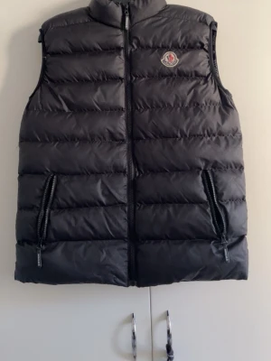 Svart dunväst från Moncler - Snygg svart dunväst från Moncler med klassisk logga på bröstet. Västen har hög krage, två dragkedjeförsedda fickor och quiltad design. Perfekt för lager-på-lager och håller dig varm med stil. Materialet är glansigt och känns exklusivt.