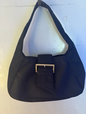 Svart handväska  - Trendig svart handväska  med brett handtag och dekorativt guldfärgat spänne framtill. Väskan har en mjuk, något kantig form och stängs med ett lock. Perfekt för dig som gillar minimalistisk och stilren design.