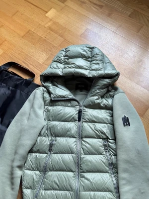 Mackage jacka  - Snygg grön dunväst från Mackage med vadderad front, två dragkedjefickor och stor huva. Ärmarna är i mjukt tyg och västen har en cool M-logga på ärmen. Perfekt för lager på lager och streetstyle.