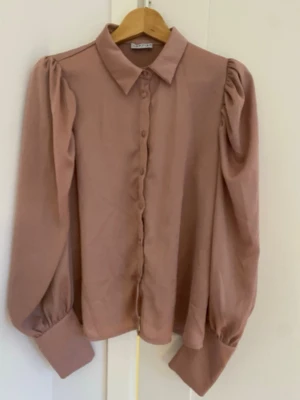 Puderrosa blus med puffärm - Trendig puderrosa blus från Lovies med puffiga långärmar och breda manschetter. Färgen är något mer rosa än vad bilderna visar. Klassisk krage och knäppning framtill. Tillverkad i ett mjukt, lätt material som ger en snygg siluett. Perfekt för dig som gillar en stilren men ändå statement look.