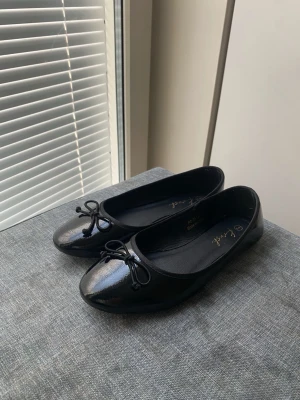 Svarta ballerinaskor med rosett - Säljer ett par klassiska, glansiga svarta ballerinaskor. Skorna har rund tå och en söt liten rosett framtill. 