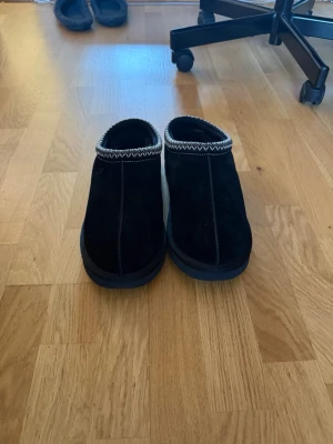 Svarta UGG tofflor i mocka - Säljer ett par svarta UGG tofflor i mjuk mocka med grov sula och dekorativt mönster i vitt runt öppningen. Perfekta för att hålla fötterna varma hemma. Klassisk slip-on modell med UGG-logga på sidan.