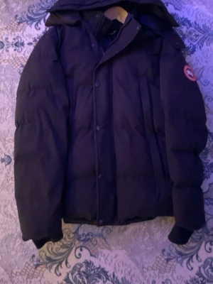 Svart dunjacka med huva Canada Goose - Säljer en svart dunjacka från Canada Goose med huva och klassisk logotyp på ärmen. Jackan har knäppning framtill, flera fickor och ribbade muddar vid ärmsluten. Perfekt för kalla vinterdagar och riktigt varm.
