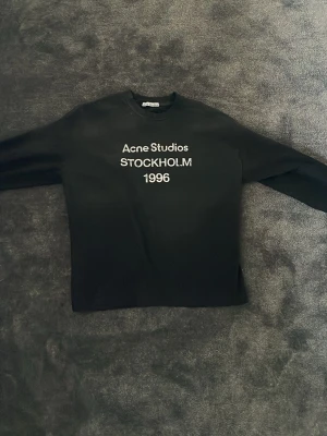 Svart sweatshirt Acne Studios - Säljer en svart sweatshirt från Acne Studios med vit text 'Acne Studios STOCKHOLM 1996' på bröstet. Tröjan har rund halsringning och långa ärmar. Perfekt för dig som gillar minimalistisk och stilren design.