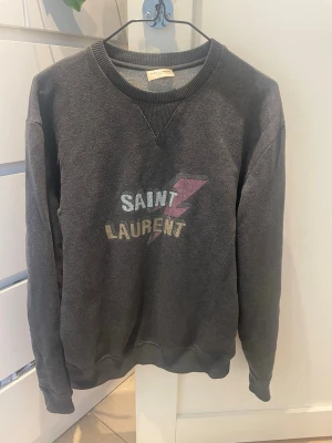  Saint Laurent sweatshirt - Cool svart sweatshirt från Saint Laurent med tryck i vitt och gult samt en rosa blixt på bröstet. Tröjan har rund halsringning med ribbade muddar vid hals, ärmslut och nederkant. Perfekt för en avslappnad och trendig look. 