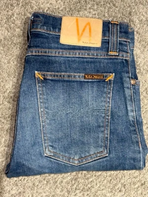 Blå jeans från Nudie Jeans - Säljer ett par klassiska blå skinny jeans från Nudie Jeans med snygga kontrastsömmar och läderpatch bak. Jeansen har fem fickor och diskret logga på bakfickan. Perfekta för dig som gillar en smal passform och tidlös denimstil.