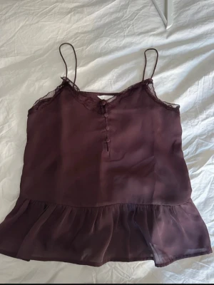 topp med spets - Säljer en vinröd linnetopp med tunna axelband och spetsdetaljer vid urringningen. Toppen har knappar framtill och ett volangparti nedtill. Perfekt för dig som gillar en feminin och luftig stil.