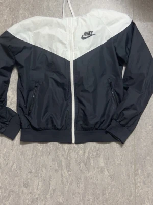 Nike svartvit vindjacka - Nike vindjacka i svart och vitt med klassisk logga på bröstet. Jackan har dragkedja framtill, två fickor med dragkedja och elastiska muddar. Tillverkad i lätt, vindtätt material och har en sportig design med V-formad färgblockning.PASSAR ÄVEN FÖR M