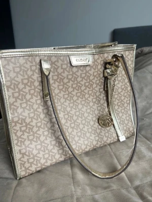 Beige vintage handväska från DKNY - Stilren beige handväska från DKNY med guldiga detaljer och logomönster över hela väskan. Väskan har två långa axelband och en dekorativ guldfärgad nyckelring med DKNY-logga. Materialet är en mix av textil och konstläder.