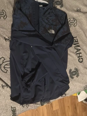 Mörkblå vindjacka från The North Face - Snygg mörkblå vindjacka från The North Face med diskret mönster upptill och logga på bröstet. Jackan har dragkedja framtill, huva och är tillverkad i ett lätt, vindtätt material som passar perfekt för blåsiga dagar.