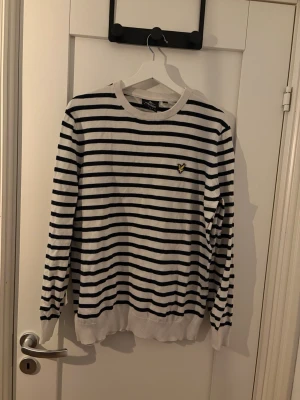 Randig tröja från Lyle & Scott - Vit långärmad tröja med svarta horisontella ränder från Lyle & Scott. Tröjan har rund halsringning och den klassiska gula loggan broderad på bröstet. Tillverkad i mjukt material och har ribbade muddar vid ärmslut och nederkant.