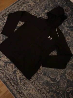 Svart hoodie från Under Armour - Säljer en svart hoodie från Under Armour med dragkedja framtill och stor logga på ryggen. Hoodien har huva, långa ärmar och en liten vit logga på bröstet. Materialet känns sportigt och passar perfekt till träning eller chill.