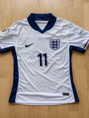 England Nike matchtröja Foden #11 - Snygg vit England matchtröja från Nike med mörkblå detaljer och nummer 11 på både fram- och baksidan. På ryggen står det 'Foden'. Tröjan har officiella märken på ärmarna och England-emblemet på bröstet. Perfekt för fotbollsfans!