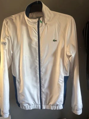 Vit vindjacka från Lacoste - Snygg vit vindjacka från Lacoste med blå detaljer längs dragkedjan och sidorna. Jackan har hög krage, elastiska muddar och diskret rutmönster på sidorna. Klassisk Lacoste-logga på bröstet och patch på ärmen. Perfekt för en sportig och stilren look.