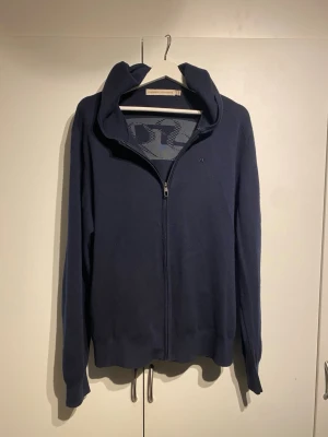 Mörkblå hoodie från J.Lindeberg merinoull - Mörkblå hoodie med dragkedja från J.Lindeberg. Stort tryck med JL-logga på ryggen och mindre logga på bröstet. Tillverkad i merinoull material, har huva och dragsko nedtill. Perfekt för en avslappnad och stilren look. Storlek L, bra skick. 