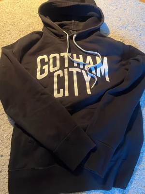 Number (N)ine Gotham City hoodie - Säljer en svart hoodie med vit text 'GOTHAM CITY' på bröstet. Hoodien har huva med vita dragsnören och en klassisk känguruficka framtill. Perfekt för dig som gillar streetwear och vill ha en bekväm och stilren look.
