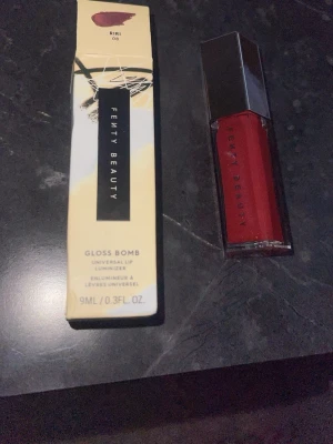 Fenty Beauty Gloss Bomb Riri 09 - Säljer ett Fenty Beauty Gloss Bomb läppglans i färgen Riri 09. Läppglanset har en djup röd nyans med glossig finish och kommer i en elegant, sexkantig plastförpackning med silvrigt lock. Innehåller 9 ml och ger läpparna en snygg glans.