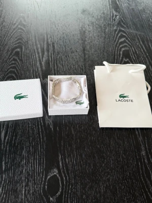 Silverfärgat armband från Lacoste - Snyggt silverfärgat armband från Lacoste med stilren och modern design. Armbandet har en länkstruktur i metall och kommer i originalförpackning med tillhörande tygpåse och presentpåse. Perfekt accessoar för dig som gillar minimalistisk och sportig stil.