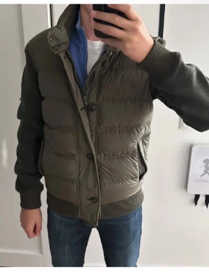 Woolrich cardigan - Snygg olivgrön pufferjacka med stickade ärmar och muddar från Woolrich. Jackan har dragkedja och knappar framtill samt hög krage. Fodrad framsida i quiltad stil och två sidofickor. Perfekt för kalla dagar och ger en sportig look. Den är i storlek s och mycket fint skick utan defekter, hör av er vid frågor eller funderingar priset går att diskutera!