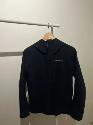 Svart vindjacka Peak Performance - Snygg svart vindjacka från Peak Performance med dragkedja framtill och två fickor med dragkedja. Jackan har huva och diskret logga på bröstet. Tillverkad i ett lätt och vindtätt material som passar perfekt för blåsiga dagar.
